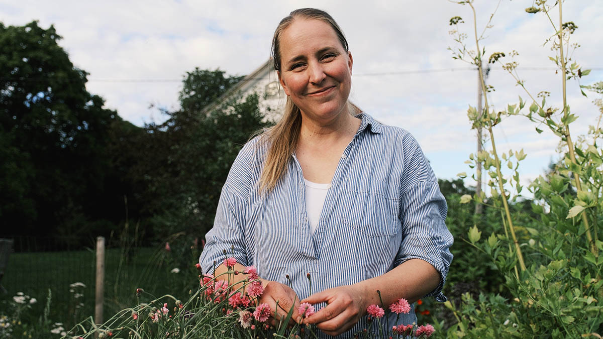 Emma Wistrand står i en trädgård och ler, klädd i en ljusblå randig tröja. Hon är omgiven av gröna växter och rosa blommor, med ett hus och träd i bakgrunden under en delvis molnig himmel.