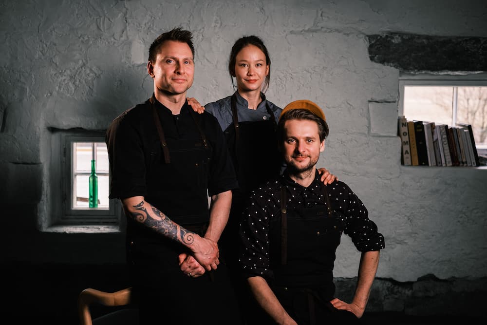 William Hellgren, Sandra Lindström and Lauri Ovaska klädda i svarta förkläden poserar tillsammans i ett rustikt rum med stenväggar, två små fönster och en hylla med böcker.