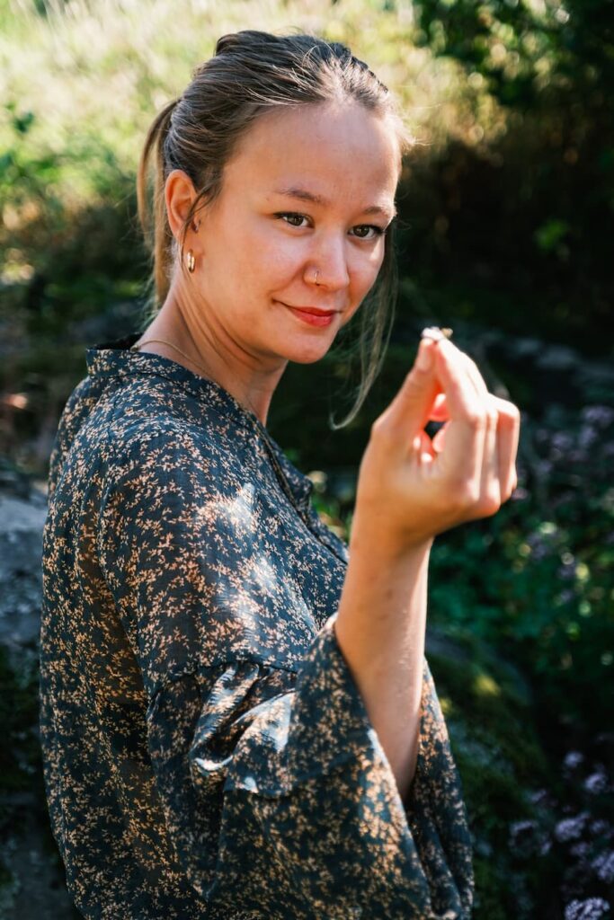 Sandra Lindström i en mönstrad blus håller upp och tittar på ett litet föremål utomhus, omgiven av grönska.