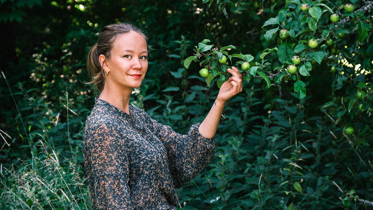 Sandra Lindström står utomhus och håller i en gren från ett äppelträd med gröna äpplen, omgiven av tätt grönt lövverk.