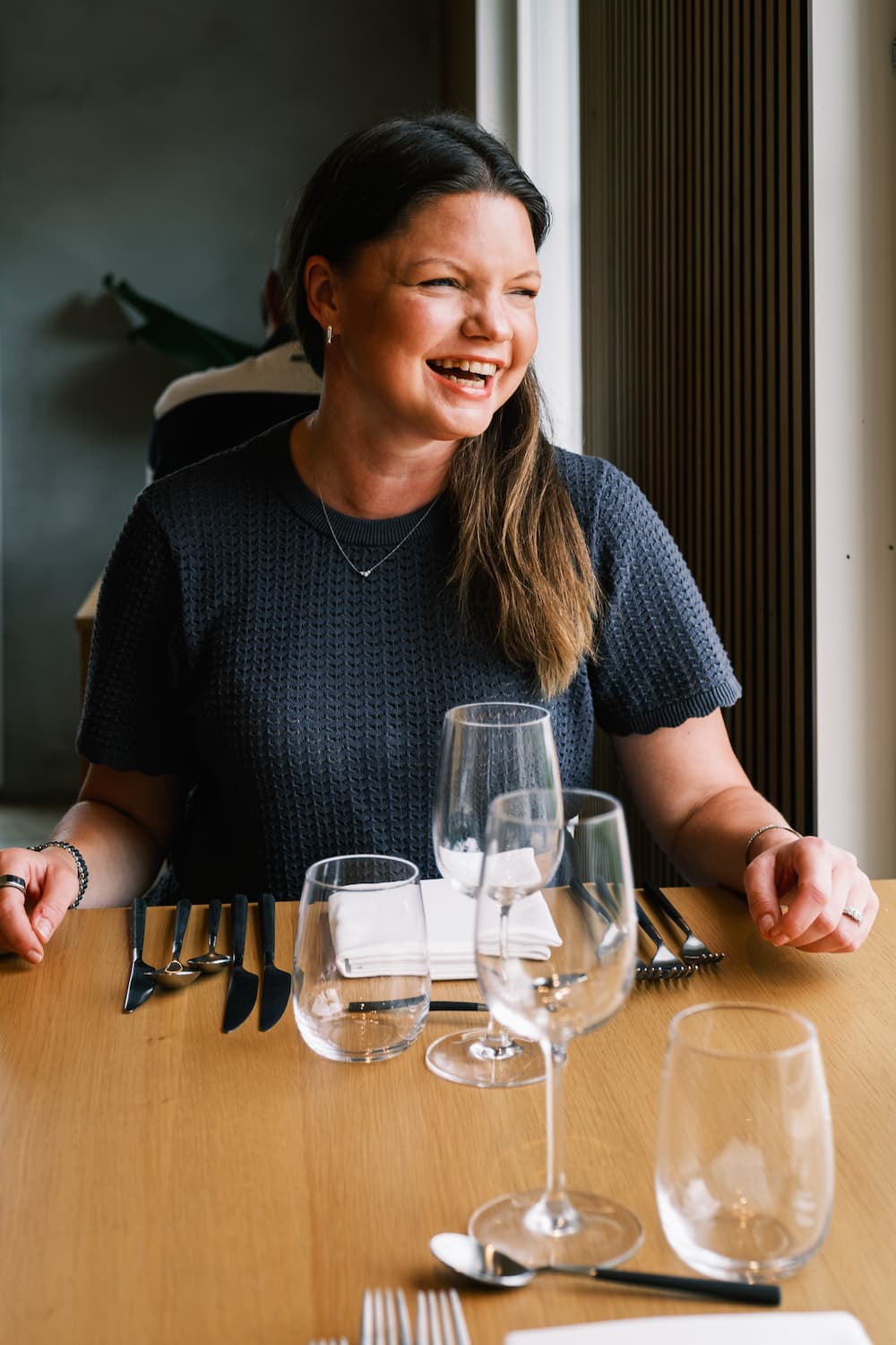 En leende kvinna sitter vid ett restaurangbord där det står vinglas och bestick.