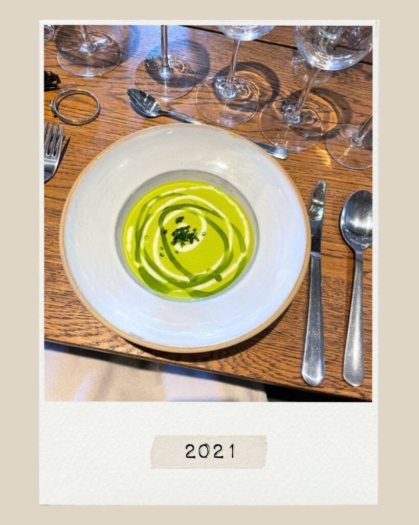 En skål med grön soppa med en krämig virvel, serverad på ett träbord med bestick och glas. Året 2021 står tryckt på en etikett längst ner.