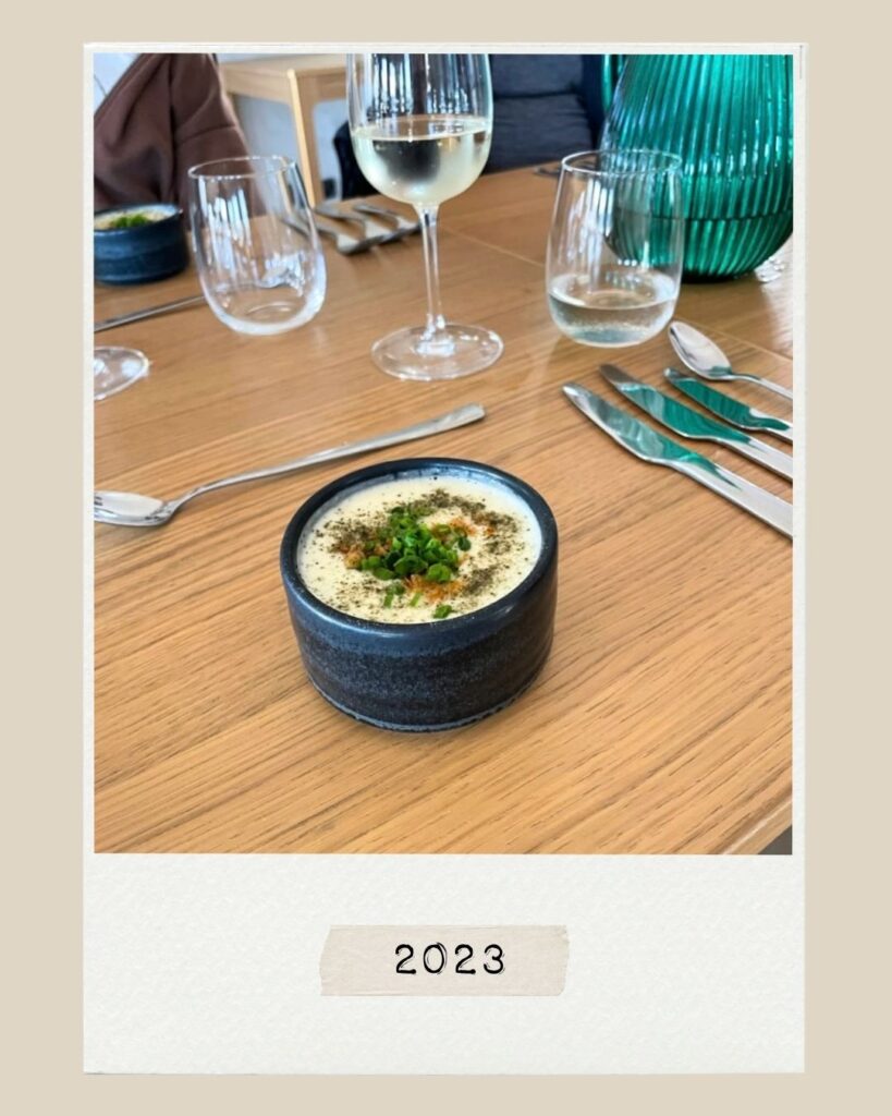 En liten skål med krämig soppa toppad med örter, på ett träbord dukat med vinglas och bestick, med en etikett där det står ”2023” nedanför.