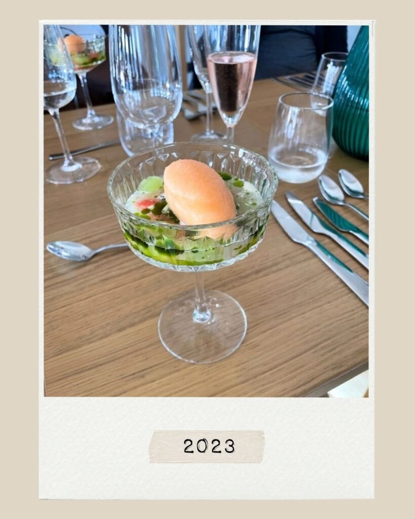 En kula apelsinsorbet serverad i ett glas med fot tillsammans med grön sås, på ett träbord med bestick och glas, märkt ”2023”.