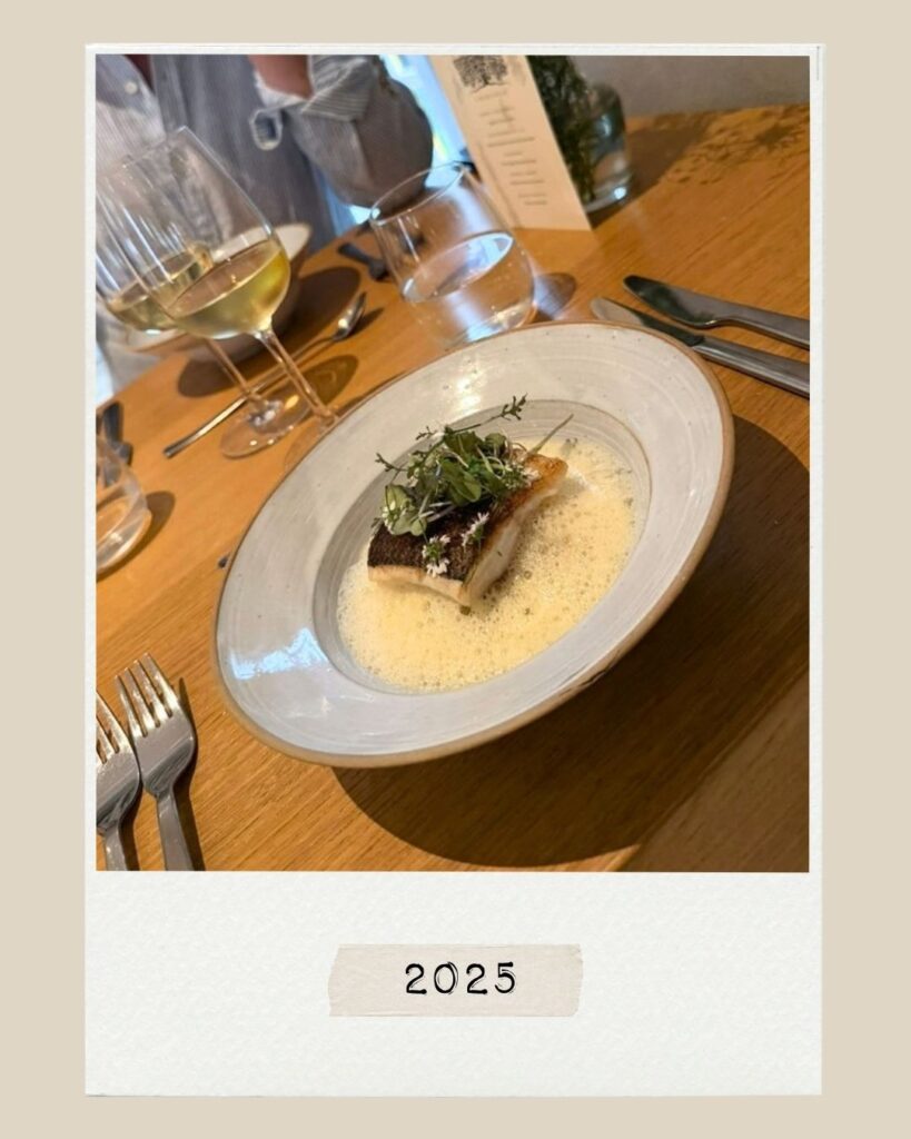 En bit stekt fisk toppad med örter i en lätt sås, serverad på ett träbord med vinglas, under texten ”2025”.
