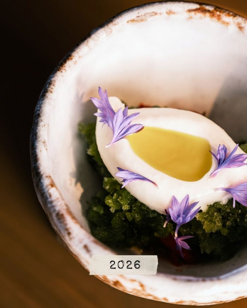 En skål med en grön örtbaserad rätt, toppad med vit grädde, gul olja och lila blomblad, med texten ”2026” på en liten etikett.